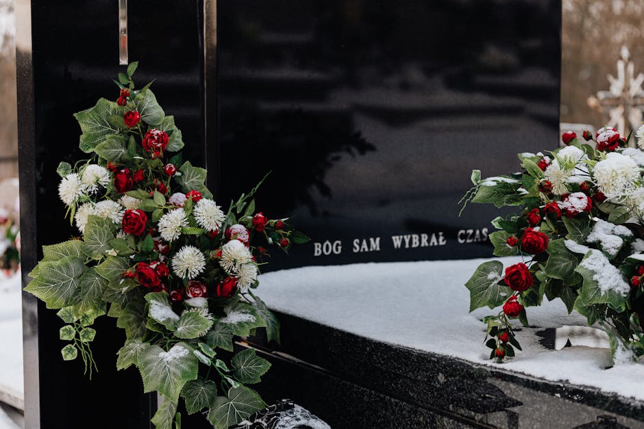 A floral tribute featuring two arrangements of red roses, white chrysanthemums, and green ivy leaves, placed on either side of a black polished stone memorial with a snow-covered surface. The flowers 