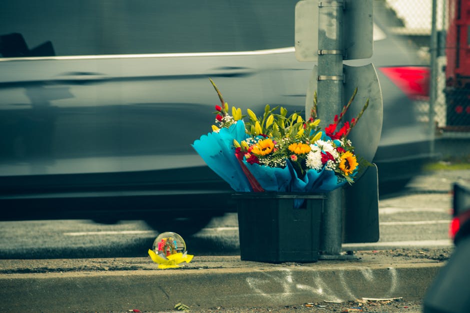 A floral tribute featuring a vibrant bouquet of fresh flowers, including yellow sunflowers, red gladioli, and white daisies, arranged with greenery and wrapped in bright blue paper. The bouquet is pla