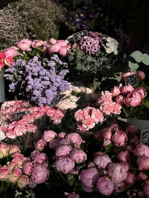 A vibrant display of fresh flowers arranged in bins and containers at floristmarksgate.co.uk. The assortment includes pink peonies with large, lush petals, clusters of lavender and purple statice, del