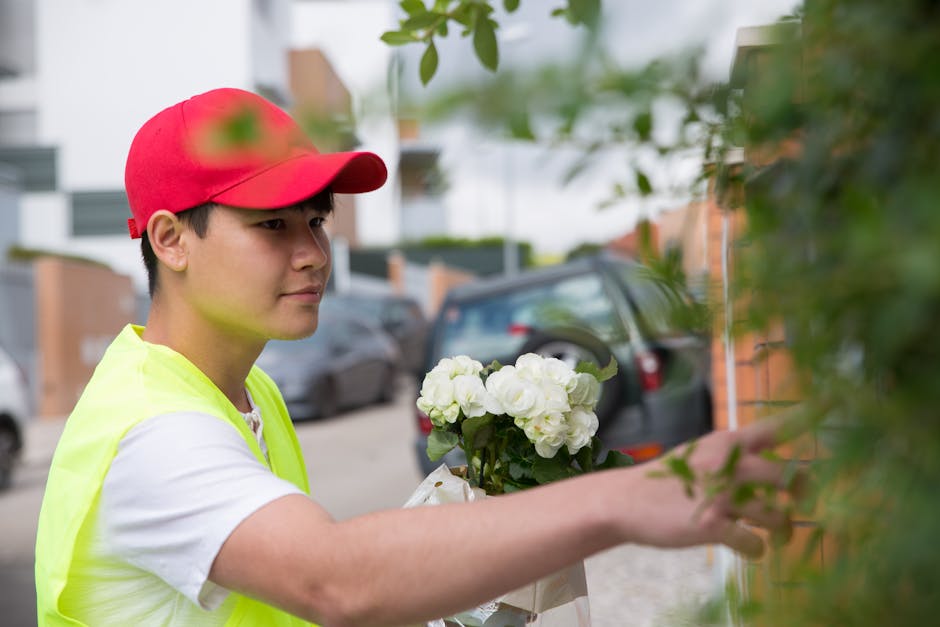 A delivery person wearing a red cap and a high-visibility yellow vest is holding a bouquet of fresh, white hydrangeas while standing outdoors on a city street. The arrangement is wrapped in white pape