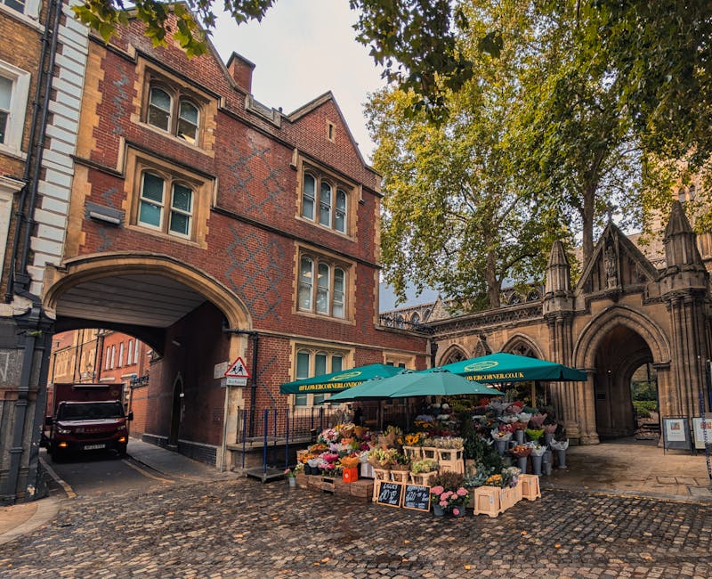 A flower stall situated on a cobbled street near Chadwell Heath station, operated by floristmarksgate.co.uk. The stall is shaded by a large green umbrella featuring the company's branding, and display