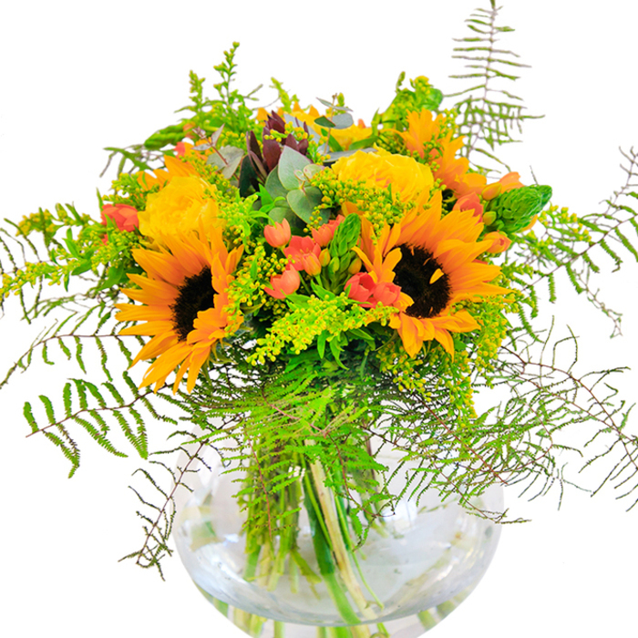 On first glance, this bouquet from Marks Gate radiates sun-kissed exuberance-a vibrant collection of golden sunflowers and sunny yellow roses blooming in harmony, their petals unfurling toward the viewer like the open arms of a warm welcome found in the neighbourhood. Each sunflower's dark brown centre adds earthy depth and instant cheerfulness, reminiscent of fields brightening the outskirts of East London. Garden roses in shades of rich yellow are woven seamlessly among the sunflowers, their tightly layered petals inviting closer admiration. Scarlet-red Hypericum berries flourish in between, their glossy skins catching the light and injecting flashes of colour that recall berry-laden hedgerows along local walkways. Fresh ferns and feathery filler plants spill from the arrangement, echoing the lushness of nearby parks and green spaces. Eucalyptus leaves emerge in their signature bluish-teal, providing gentle aroma and a hush of tranquillity. The bouquet is artfully held in a clear, bowl-shaped vase that reveals the vibrant tapestry of flower stems basked in clean water. Suitable for birthdays, anniversaries, or spontaneous acts of kindness, this creation from a skilled Marks Gate florist brings together the seasonal bounty and heartfelt hospitality of the area-an arrangement made to light up any occasion.